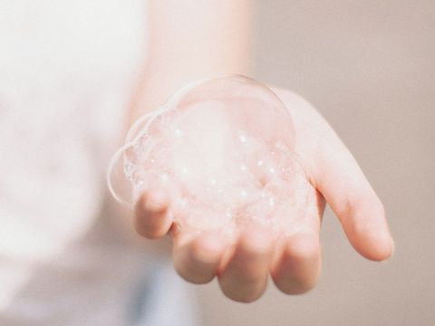 La mano di una persona tiene un insieme di bolle di sapone delicate e traslucide contro uno sfondo morbido e sfocato. Le bolle appaiono iridescenti con accenni di riflessi di luce.