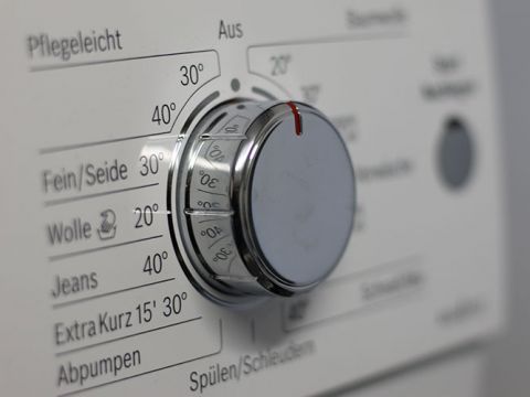 Primo piano del selettore di una lavatrice con varie impostazioni, tra cui "Delicati/Seta", "Lana", "Jeans" e "Extra breve 15' 30°". Il selettore è impostato su 30° vicino a "Lana". Testo tedesco etichetta vari programmi di lavaggio e temperature.