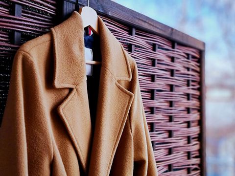 Un cappotto color cammello è appeso a una gruccia bianca davanti a un paravento di legno intrecciato con uno sfondo sfocato. Il cappotto presenta un motivo a revers.