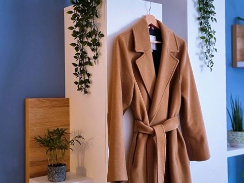 Un cappotto marrone è appeso a un gancio su una parete bianca, affiancato da piccole piante verdi. La parete e l'ambiente circostante sono decorati con ancora più verde, creando un'atmosfera fresca e naturale.