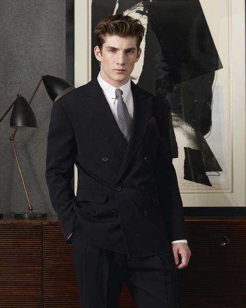 Un uomo in doppiopetto nero e cravatta grigia è in piedi con sicurezza di fronte a un'opera d'arte astratta. Accanto a lui, su una superficie di legno, c'è una lampada nera con dettagli in ottone. L'ambiente è moderno ed elegante.