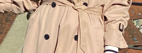 Una persona indossa un trench doppiopetto beige con cintura in vita e si trova di fronte a uno sfondo di tetti. Il cappotto ha bottoni scuri e polsini regolabili.