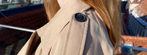 Una persona con i capelli lunghi indossa un trench beige su un tetto. Il focus è sulla spalla del cappotto con un bottone decorativo. Sullo sfondo si vedono tetti e un cielo sereno.