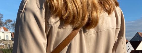 Una persona con lunghi capelli castano chiaro che distoglie lo sguardo, indossa una camicia beige e una tracolla marrone. Si trova all'aperto con un cielo azzurro limpido e una vista parziale di edifici e alberi sullo sfondo.