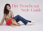 Eine Person mit langen, dunklen Haaren, die einen beigen Trenchcoat, blaue Jeans und weiße Turnschuhe trägt, sitzt auf dem Boden. Sie hat eine rote Handtasche dabei und lächelt. In roter Schrift ist der Text „Der Trenchcoat-Style-Guide“ zu sehen.