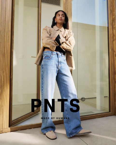Una donna è in piedi con sicurezza davanti a una porta di vetro, indossa una giacca beige, un top nero, jeans larghi blu e scarpe beige. Il testo "PNTS MADE BY HUMANS" è visualizzato nella parte inferiore dell'immagine.