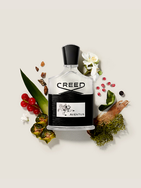 Una bottiglia di profumo Creed Aventus circondata dai suoi ingredienti: bacche rosse, fiori bianchi, ananas, muschio e alcune spezie scure. Lo sfondo è in un tenue beige.