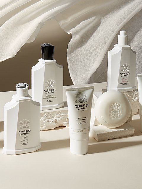 Una presentazione lussuosa di prodotti per la cura della pelle e profumi Creed, tra cui flaconi, un tubetto e un pezzo di sapone. Tutti gli articoli sono disposti elegantemente su una superficie dai toni neutri con un tessuto morbido e fluente sullo sfondo.
