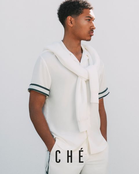 Un uomo in abito bianco con finiture nere, composto da camicia a maniche corte e pantaloni, posa davanti a un semplice sfondo bianco. Un maglione bianco è appoggiato sulle spalle, le mani sono in tasca. In basso c'è il testo "CHÉ".