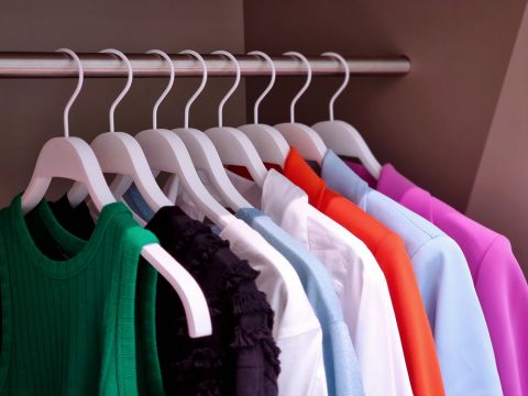 Una serie di capi d'abbigliamento colorati su appendini bianchi sono ordinatamente disposti su una barra di metallo. I capi sono disponibili in vari colori come verde, nero, bianco, grigio, blu, arancione e rosa, offrendo una presentazione vivace e ordinata.