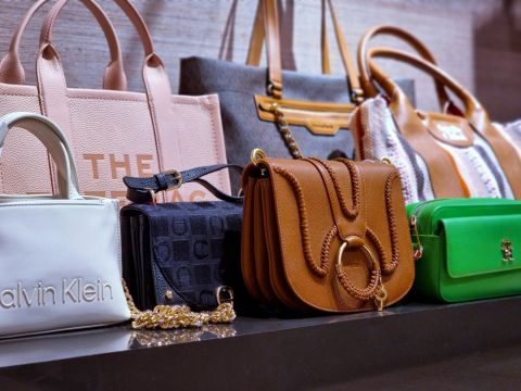 Una mostra di diverse borse di design, tra cui una borsa bianca di Calvin Klein, una borsa in pelle marrone con tracolla a catena, una pochette verde e diverse borse tote in vari colori e fantasie, disposte su uno scaffale.