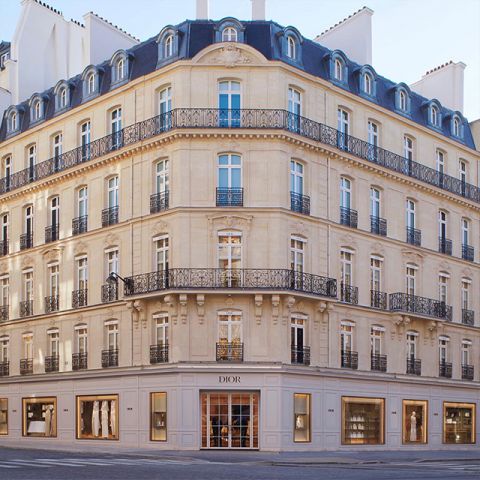 Elegante edificio angolare con architettura classica, diversi piani, finestre ad arco e balconi in ferro battuto decorati. Al piano terra si trova un negozio Dior con ampie vetrine. La luce naturale intensa esalta la facciata beige.