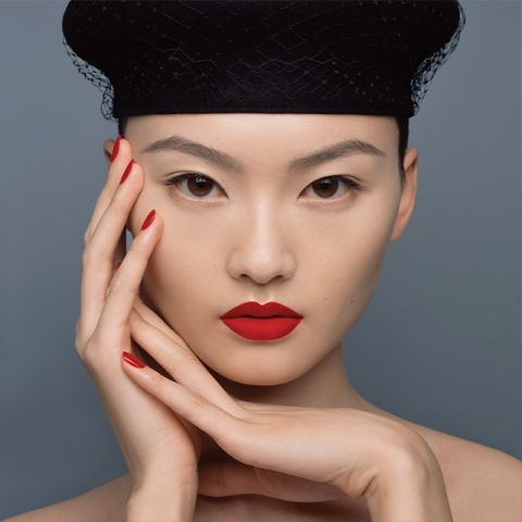 Una persona con rossetto e smalto rosso acceso posa su uno sfondo grigio. Indossa un cappello nero con una struttura a rete e le sue mani sono elegantemente posizionate intorno al viso, enfatizzando il trucco vivace.