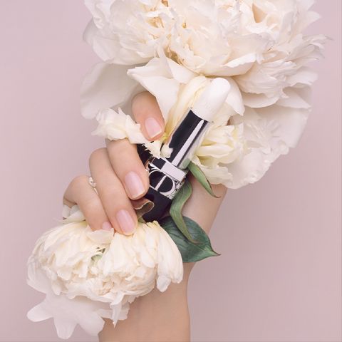 Una mano con unghie curate tiene un rossetto bianco, circondata da grandi fiori di peonia bianca, creando un'atmosfera delicata ed elegante. Lo sfondo è rosa chiaro e completa l'estetica delicata.