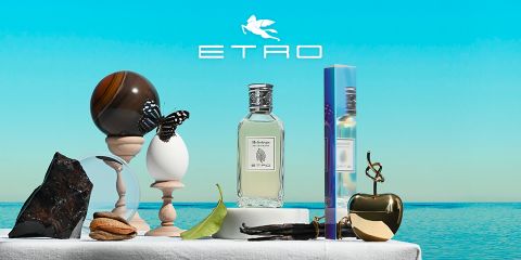 Flacone di profumo con la scritta "ETRO" e un design a pigna, circondato da decorazioni artistiche: una sfera, un uovo con una farfalla, una pietra, una foglia, noci, una mela dorata e una mano scolpita. In alto, il logo ETRO su uno sfondo di cielo blu e mare.