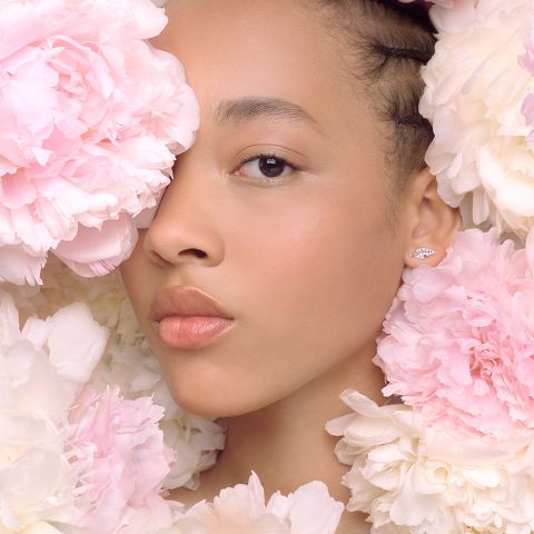 Una donna è parzialmente circondata da grandi peonie rosa e bianche. Il suo viso è delicatamente illuminato, mostrando un trucco naturale e labbra lucide. All'orecchio visibile indossa un piccolo ed elegante orecchino. Il tono generale dell'immagine è dolce e sereno.
