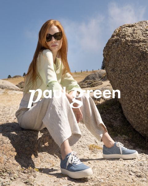 Una persona con lunghi capelli rossi, occhiali da sole, un top verde chiaro, pantaloni beige e scarpe da ginnastica blu è seduta su un terreno roccioso sotto un cielo azzurro. Le parole "Paul Green" sono sovrapposte all'immagine.