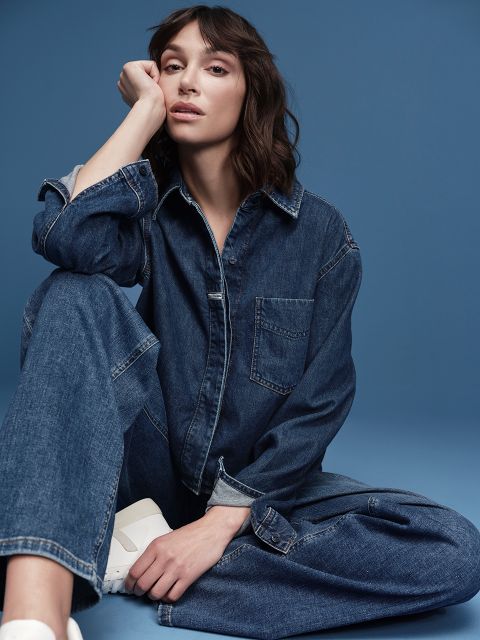Una persona con capelli castani lunghi fino alle spalle è seduta sul pavimento davanti a uno sfondo blu. Indossa una salopette di jeans e scarpe da ginnastica bianche. Si appoggia il mento sulla mano e sembra rilassata.