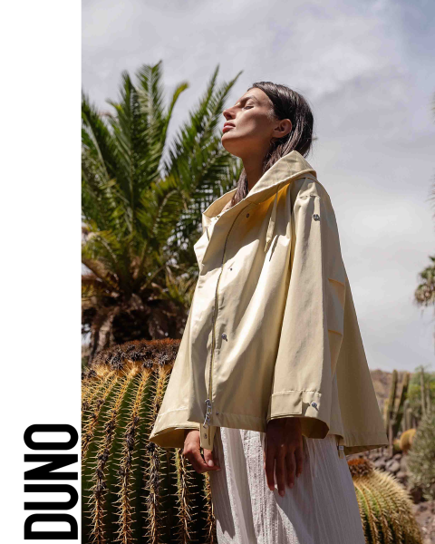 Una persona è di profilo, indossa una giacca giallo chiaro e una gonna bianca, ha la testa reclinata all'indietro e gli occhi chiusi. Si trova in un ambiente esterno con grandi cactus e palme. La parola "DUNO" appare verticalmente sul lato sinistro dell'immagine.