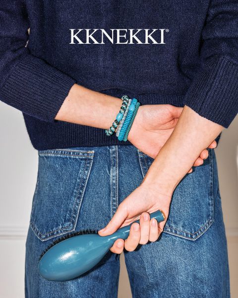 Una persona con un maglione blu navy e jeans tiene una spazzola per capelli blu dietro la schiena e indossa braccialetti di perline turchesi. Sopra il logo "KKNEKKI".