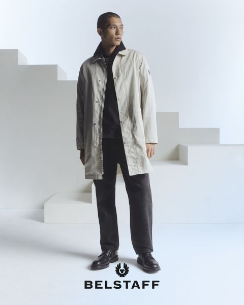 Una persona che indossa un cappotto chiaro sopra pantaloni e scarpe scure è in piedi di fronte a uno sfondo minimalista e geometrico. Nella parte inferiore dell'immagine è visibile il logo "Belstaff".