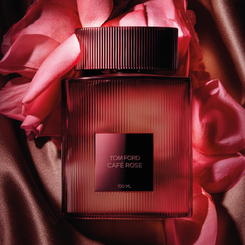Una bottiglia del profumo Tom Ford Café Rose è circondata da petali di rosa rosa su un tessuto setoso color bronzo. La bottiglia è rettangolare con scanalature verticali e un'etichetta scura e quadrata al centro, che trasmette lusso ed eleganza.