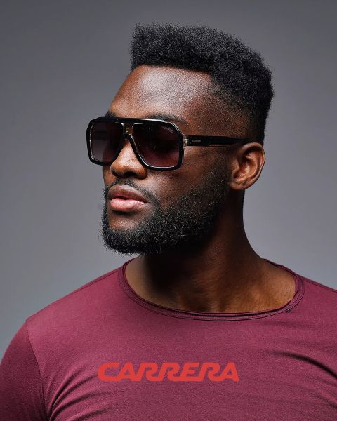 Uomo che indossa grandi occhiali da sole quadrati e una camicia color vinaccia con il logo "CARRERA", guarda di lato, su uno sfondo grigio.