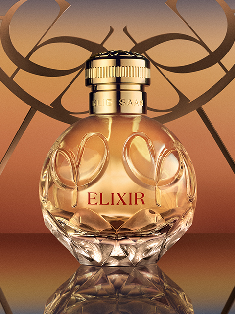 Un flacone di profumo in vetro con la scritta "Elie Saab Elixir" e un tappo dorato si trova davanti a uno sfondo caldo con sfumature. Il flacone rotondo è decorato con incisioni floreali decorative e riflette la luce, conferendogli un aspetto elegante e lussuoso.