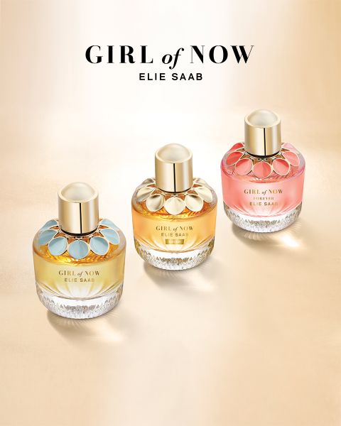 Tre flaconi di profumo "Girl of Now" di Elie Saab sono presentati su una superficie oro chiaro, ognuno con una combinazione di colori unica: accenti blu, oro e rosa. I tappi sono dorati e il nome del marchio con il titolo della collezione è posizionato sopra i flaconi.