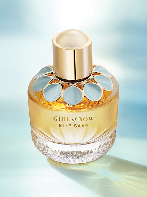 Una bottiglia di vetro di "Girl of Now" di Elie Saab è posizionata di fronte a uno sfondo morbido ed etereo. La bottiglia presenta accenti dorati e un motivo floreale attorno al collo. La fragranza contiene un liquido ambrato caldo, che contribuisce a un aspetto lussuoso ed elegante.
