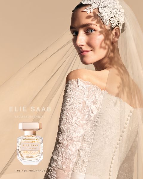 Una sposa in abito di pizzo e velo, con un accessorio floreale per capelli, guarda oltre la sua spalla. Il suo abito proietta un'ombra morbida con un motivo floreale su uno sfondo beige. È esposta una bottiglia di profumo con il testo "Elie Saab Le Parfum Bridal".