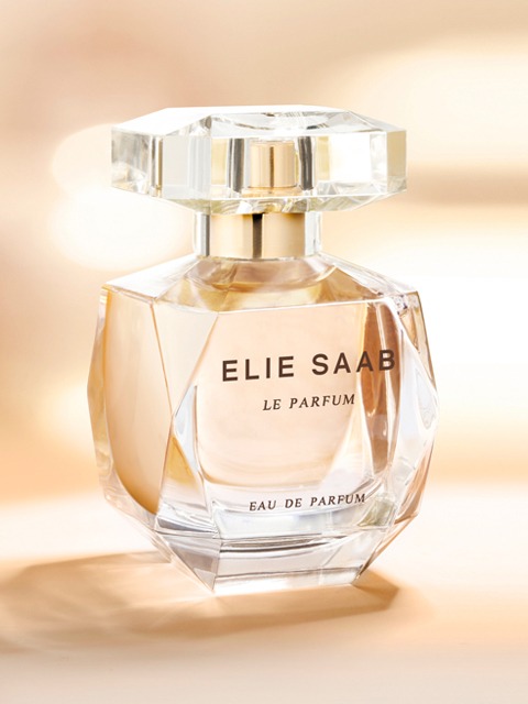 Su uno sfondo morbido e sfocato, si vede un lussuoso flacone di profumo con la scritta "Elie Saab Le Parfum". Il flacone è elegante, in vetro trasparente con un design geometrico e sfaccettato e contiene un liquido dorato.