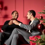 Zwei Personen sitzen auf einem roten Sofa in einem festlich geschmückten Raum. Sie lächeln und stoßen an. Im Vordergrund sind ein Weihnachtsbaum und rote Weihnachtskugeln zu sehen. Eine Person trägt ein schwarzes Oberteil, die andere einen grauen Anzug.