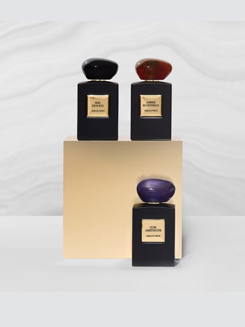 Tre eleganti flaconi di profumo su un piedistallo beige. Ogni flacone è rettangolare e ha un tappo ovale unico: uno nero, uno rosso e uno viola. Lo sfondo presenta un motivo ondulato sottile.