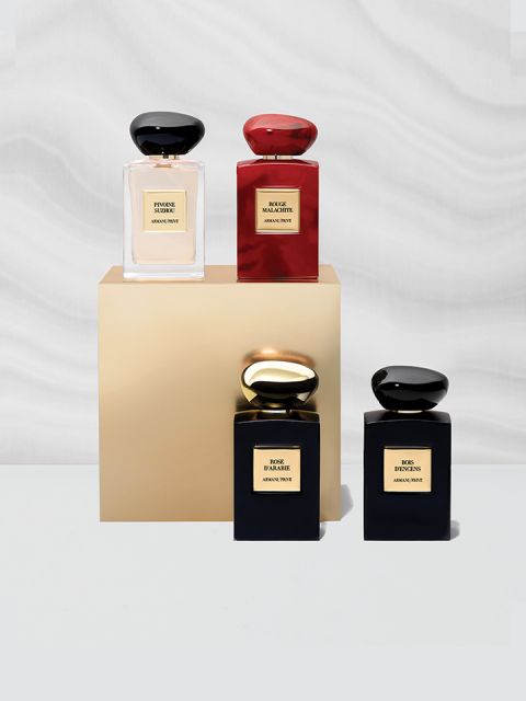 Quattro eleganti flaconi di profumo esposti su una base dorata. I flaconi sono neri, rossi e dorati, ciascuno con un tappo abbinato. Sono disposti su uno sfondo morbido, ondulato, grigio-bianco.
