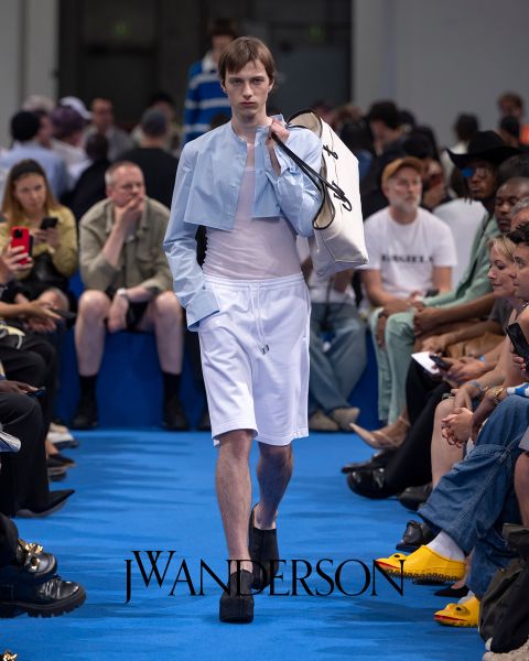 Una modella su una passerella blu indossa una maglietta corta azzurra, pantaloncini bianchi e scarpe platform nere e porta una grande borsa bianca. Gli spettatori seduti guardano e scattano foto. Il logo di J.W. Anderson è visibile sul pavimento.