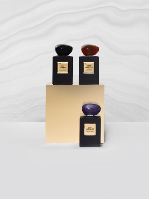 Tre flaconi di profumo su una base dorata davanti a uno sfondo bianco ondulato e strutturato. I flaconi sono neri con etichette dorate e tappi ovali lisci e scuri.