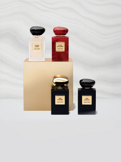 Quattro flaconi di profumo sono esposti sopra e intorno a un piedistallo beige. I flaconi sono quadrati con tappi arrotondati e sono nei colori nero e rosso. Una parete bianca ondulata e strutturata fa da sfondo.