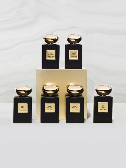 Sono esposti sei eleganti flaconi di profumo con lisci tappi dorati. Sono disposti sopra e intorno a un blocco dorato e presentano varie etichette con le scritte "OUD", "SNAKE" e "OUR", ciascuna con elaborati motivi neri e dorati su un delicato sfondo grigio.