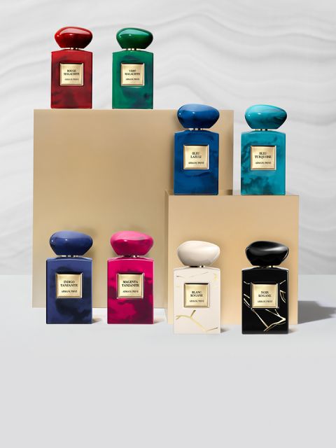 Otto flaconi di profumo in vari colori, tra cui rosso, verde, blu, rosa e nero, sono disposti su piedistalli beige davanti a uno sfondo chiaro. Ogni flacone ha un'etichetta con scritte dorate.