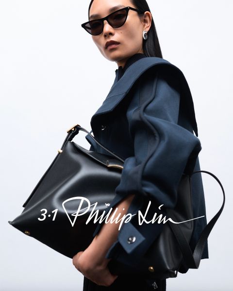 Persona con occhiali da sole, cappotto scuro e una grande borsa nera su uno sfondo semplice. La sovrapposizione di testo recita in una calligrafia stilizzata "3.1 Phillip Lim".