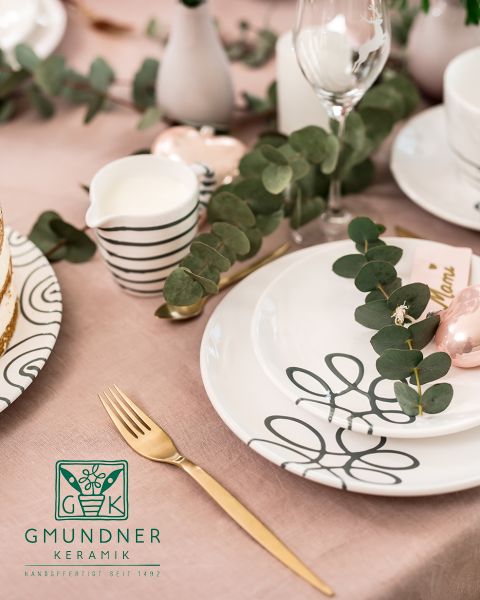 Una tavola splendidamente apparecchiata con piatti decorativi in ceramica con motivi astratti a linee nere. Foglie di eucalipto adagiate sui piatti. Una forchetta dorata, una lattiera a righe e una tazza decorata completano l'elegante tovaglia rosa.
