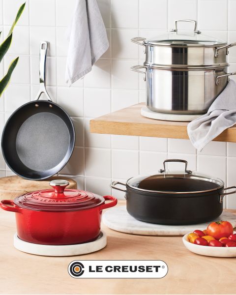 Una scena di cucina con una casseruola rossa, una padella antiaderente, un set di pentole impilabili in acciaio inossidabile, una pentola nera e una ciotola di pomodorini su un bancone di legno chiaro davanti a un paraschizzi di piastrelle bianche. Il logo Le Creuset è visibile.