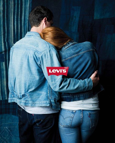 Uomo e donna si abbracciano davanti a uno sfondo patchwork blu, entrambi indossano giacche di jeans e jeans. Sopra di loro spicca chiaramente un logo Levi's rosso.