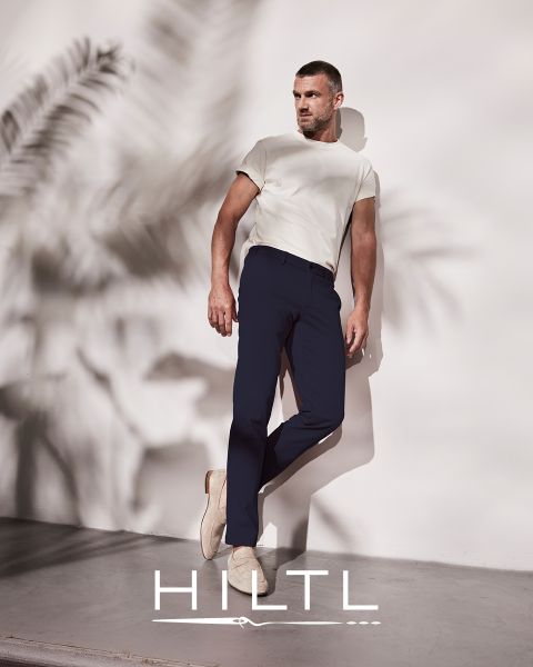 Un uomo in maglietta bianca e pantaloni blu scuro è appoggiato a un muro con foglie di palma. Indossa scarpe beige. La scena è minimalista. Nella parte inferiore dell'immagine c'è la parola "HILTL".