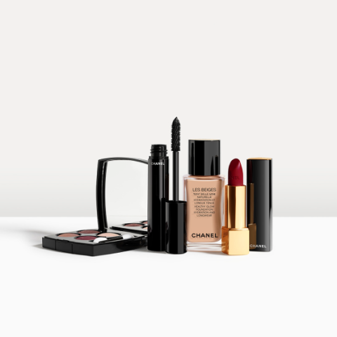 Una collezione di prodotti per il trucco di Chanel, tra cui un set compatto di ombretti, un mascara, un fondotinta liquido in un flacone di vetro e un rossetto rosso scuro in un astuccio oro e nero, disposti su una superficie bianca.