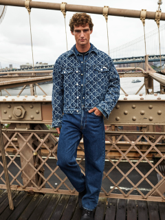 Un uomo con una giacca di jeans fantasia e jeans blu è in piedi su un ponte con ringhiera metallica. Sullo sfondo si vedono un fiume e un altro ponte in una giornata nuvolosa.