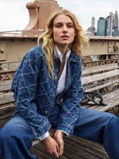 Una persona con lunghi capelli biondi è seduta su una panchina di legno e indossa una giacca di jeans blu, una camicia bianca e jeans larghi. Sullo sfondo si vedono edifici urbani e grattacieli.