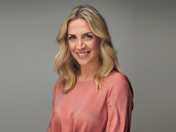 Una persona con lunghi capelli biondi ondulati e un sorriso caloroso indossa un top a maniche lunghe color corallo. Lo sfondo è in un grigio neutro, conferendo all'immagine un aspetto professionale e amichevole.