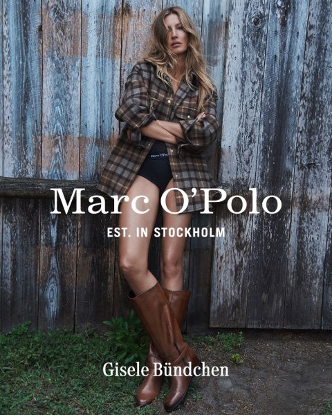 Una donna con una camicia a quadri, pantaloncini neri e stivali marroni è in piedi davanti a una parete di legno consumata. Il testo recita: "Marc O'Polo EST. IN STOCKHOLM Gisele Bündchen.